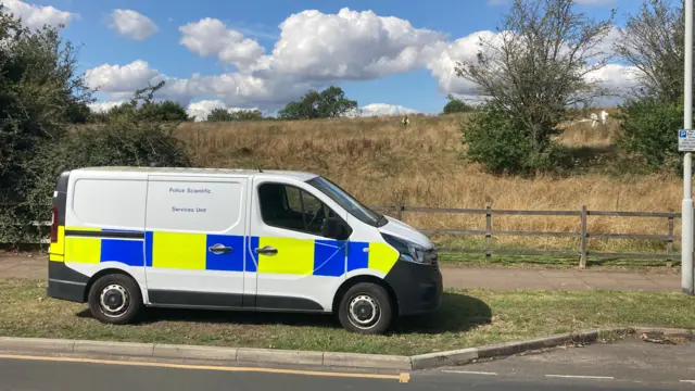 A police van is parked in front of a field where forensic investigators and police officers are working.