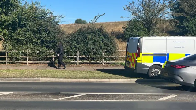 An armed officer is walking on the pavement alongside a field. A Police Scientific Services Unit van is nearby