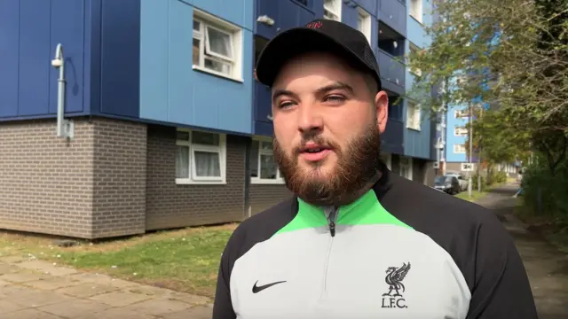 Davy Bridgestock, wearing an LFC sports top and a hat, standing in front of the blue cladding of Leabank