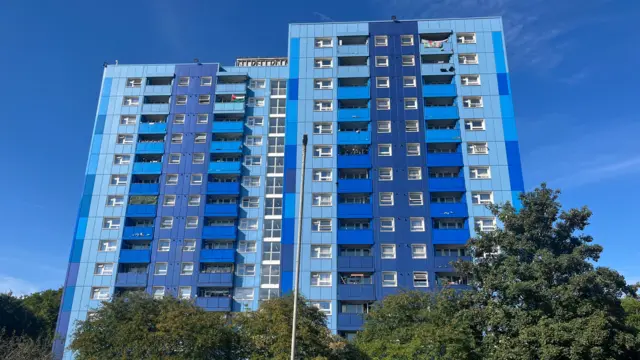 A tall blue tower block on a clear day. Green trees sit at the foot of it.