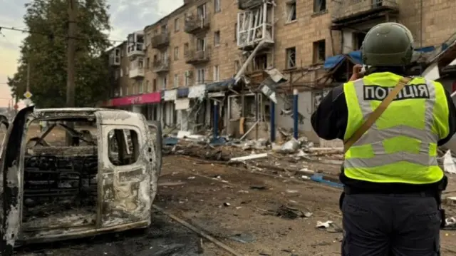 A person in a high-vis vest stands next to a burned-out van, taking a photo of a damaged building