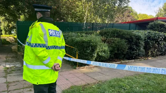 An officer wearing a high vis jacket stands near a piece of police tape near the block of flats.