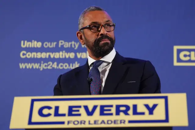 James Cleverly in suit with blue and red tie behind a 'Cleverly for leader' branded podium