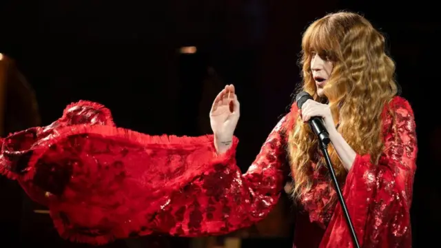 Florence + The Machine singing at the Proms. She is wearing a red dress with large, flowy sleeves while holding a microphone on a microphone stand