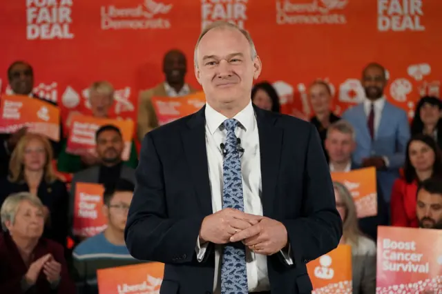 Sir Ed Davey stands in the foreground while in the background, people stand with orange Liberal Democrat placards