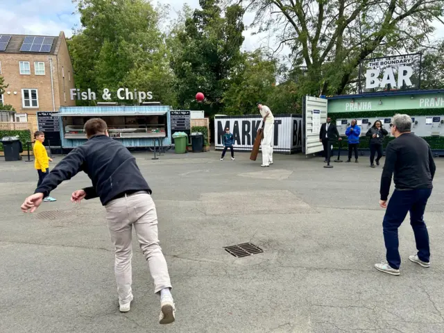 Fans play cricket with players on stilts outside the ground