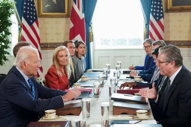 US President Joe Biden and UK Prime Minister Sir Keir Starmer sit at a long table with files, glasses of water, and tea cups on it. Other people are also sat at the table.