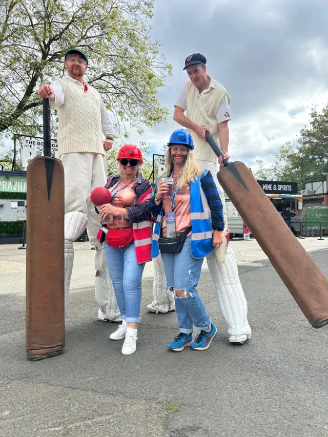 Performers on stilts dressed as cricketers pose for a picture with two women wearing high vis jackets and hard hats