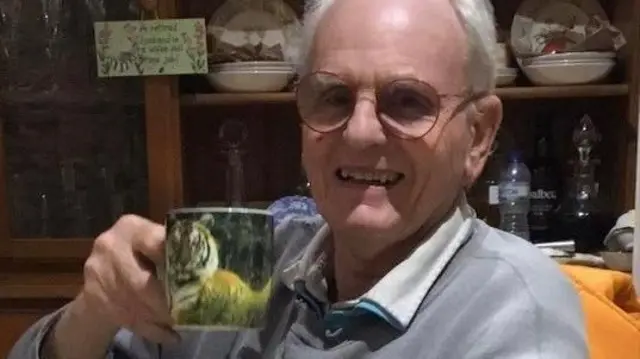 Pensioner Jon Harvey is wearing glasses and a grey jumper while smiling at the camera and holding a mug with a picture of a tiger on it