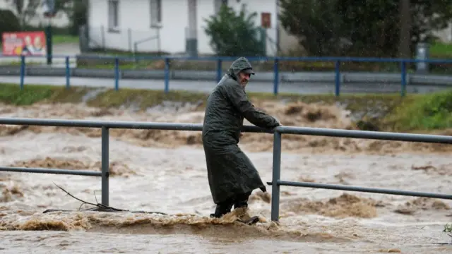 A man wearing a full ternch coat holds onto a railing on a flood-affected road