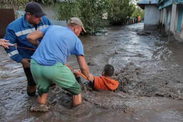 A man wearing an orange hoody being pulled out of the water by two men