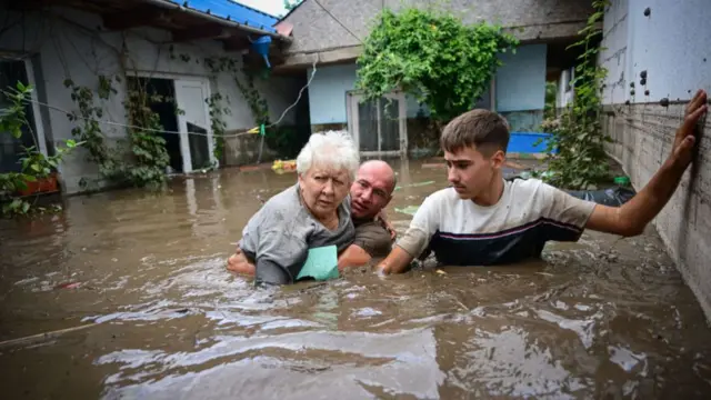 Two men rescue an elderly woman. Brown flood water reaches their chests. A building is behind them.