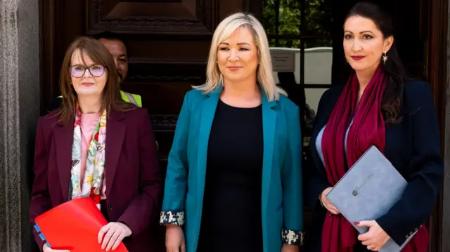 Archibald is wearing a maroon blazer, flowery pink, yellow, green and yellow scarf and a pink top, O'Neill is wearing a turquoise blazer and black top and Little-Pengally is wearing a maroon scarf, navy blazer. The ministers are standing outside an official looking building with a security guard in vision. Archibald is holding a red file and Little Pengelly is holding a blue file.