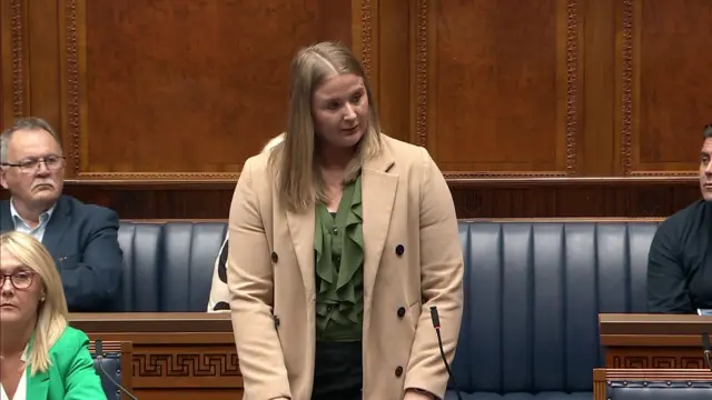 A woman with blond hair stands in the Assembly chamber. She is wearing a light coloured coat and a green top. She is standing in front of blue leather benches