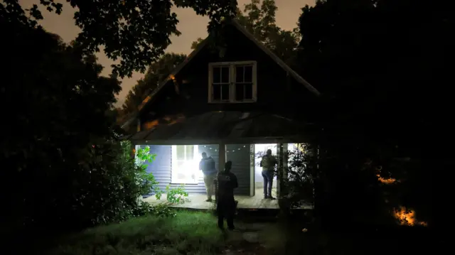 A suburban house at night, illuminated by torchlight as three people stand outside