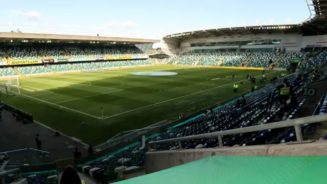 A wide view of the inside of Windsor Park. There are green and white plastic chairs in the stands and a grass pitch with football markings.