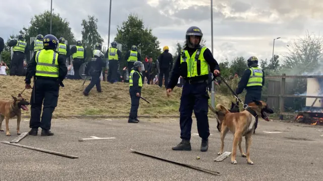 Police dog handlers in riot gear hold on to their animals as the ground has shards of broken fencing panels strewn around