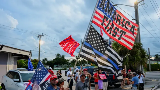 People on the sidewalk waving flags that say "AMERICA FIRST", some of them wearing MAGA hats