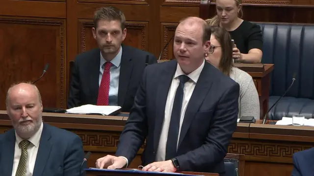 A man in a navy suit and tie stands at a wooden podium in the Assembly chamber