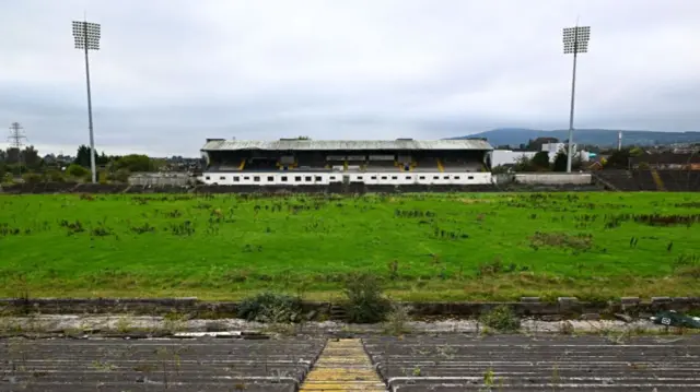 A wide image of an overgrown grass stadium. In the distance you can see concrete stands with flood lights either side,