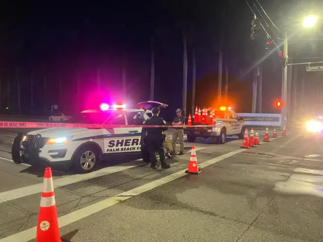 Four police officers stand next to a police car with its lights on, alongside a long string of police tape and traffic cones