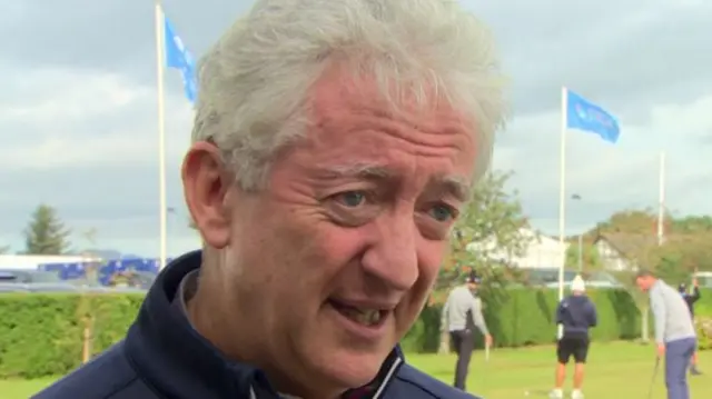 John has grey hair and blue eyes and has blue flags behind him and golfers by a flag.