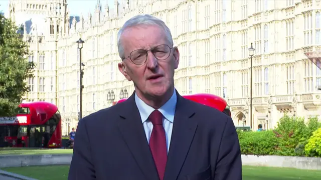 A man in a dark navy suit stands in front of parliament buildings. He is wearing a red tie and talking to the camera.