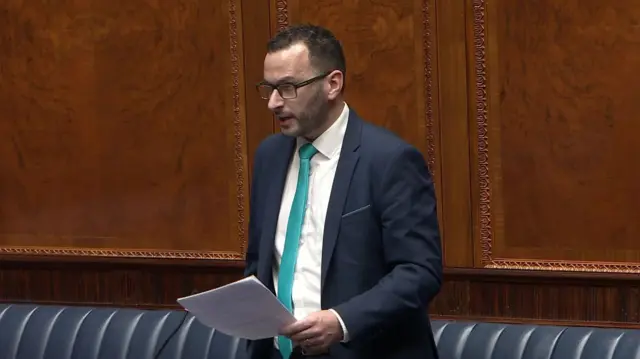 A man in a navy suit and white shirt stands in the Assembly chamber. He is wearing dark-rimmed glasses and a light blue tie and is holding a piece of paper.