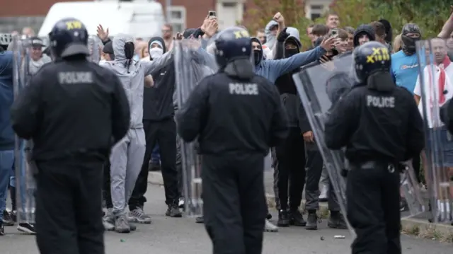 Masked men gesticulate to riot police officers who are wearing helmets and are behind a wall of transparent shields