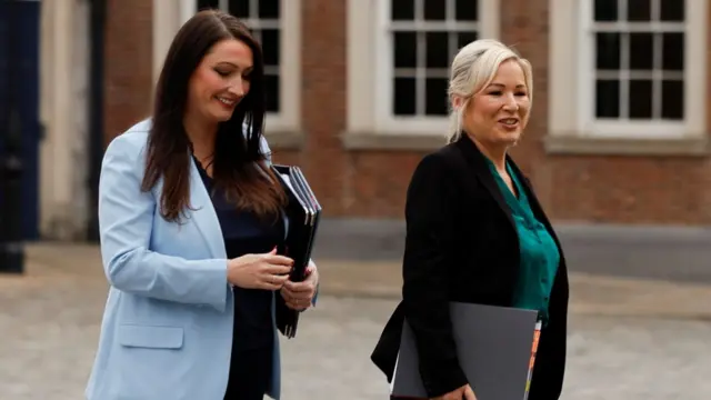 Emma is wearing a blue blazer and black top. She has long brown hair and is smiling at the ground. She is holding a briefcase. Michelle O'Neill is smiling ahead. She is wearing a green V-neck top and black blazer. She has blond hair that is tied up with her fringe down to one side. In the background, there are white windows and red bricks.