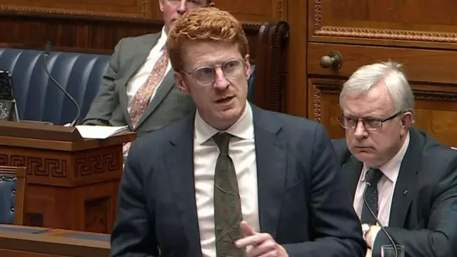 Matthew wearing a green tie and white shirt and navy suit. He has glasses and red hair. He is in the chamber.