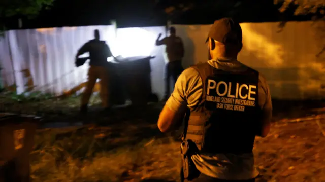 Three men, one wearing a police vest, stand near a fence illuminated by torchlight