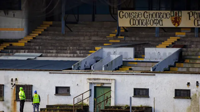 Two workers in his visibility jackets look up at a concrete terraced stand.