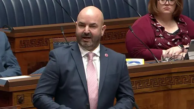 A man in a grey suit and pink tie stands on wooden benches in the NI Assembly