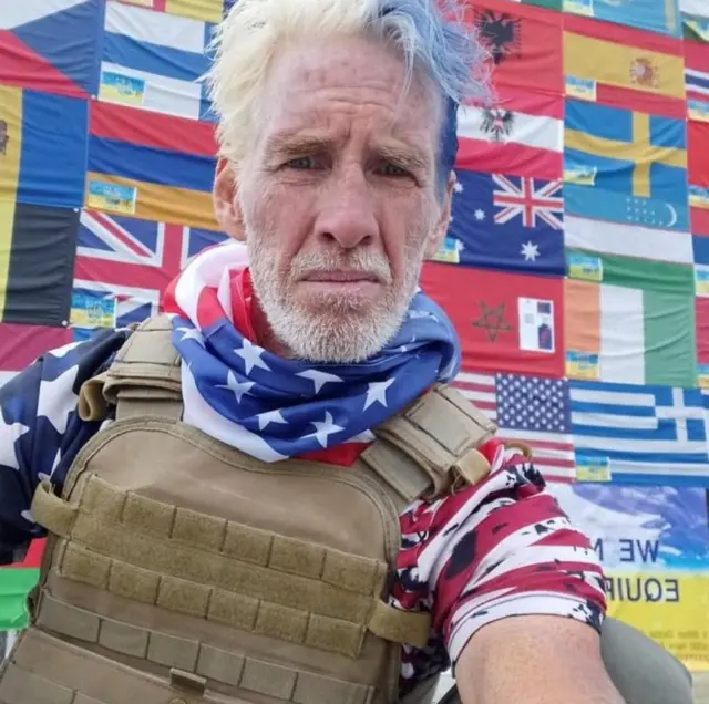 An undated selfie shows Ryan W. Routh in a military bulletproof vest wearing a red white and blue t-shirt and bandana around his neck with a wall of flags of the world behind him