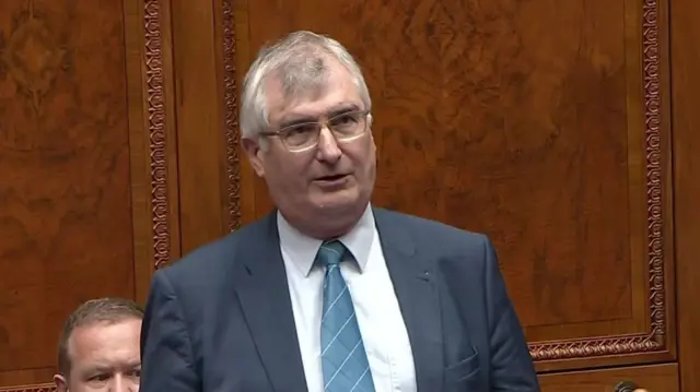 A man with a grey suit and blue tie standing in front of a wooden panel