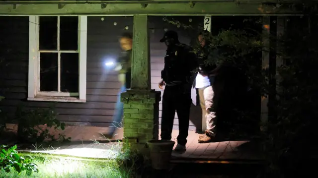 Three people, two with torches, on the front porch of a suburban house at night