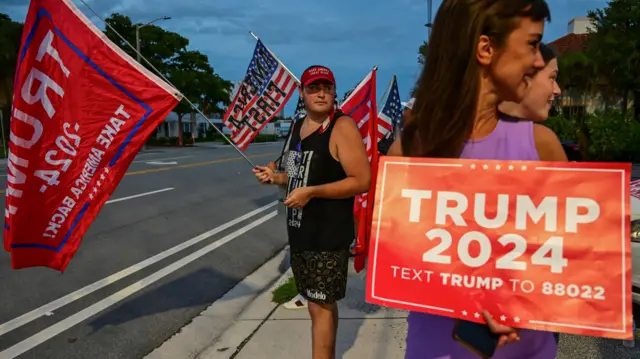 People standing in the street with signs and flags saying "TRUMP 2024"