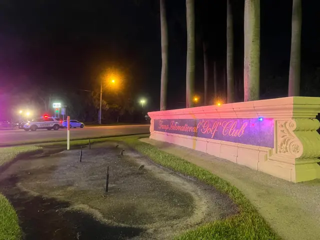 A large plaque signposting the Trump International Golf Club, with police cars in the background