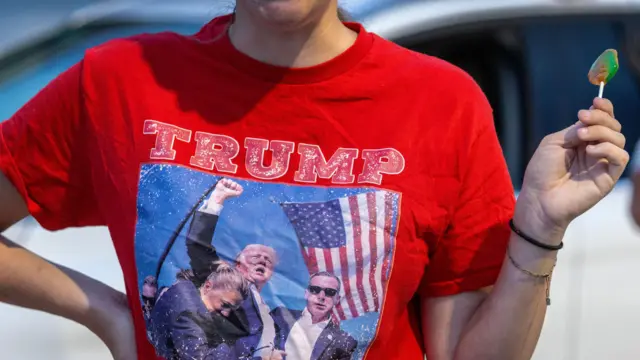 Person holding a lollipop and wearing a red t-shirt that says 'TRUMP BULLETPROOF' alongside an image of Donald Trump after surviving a shooting