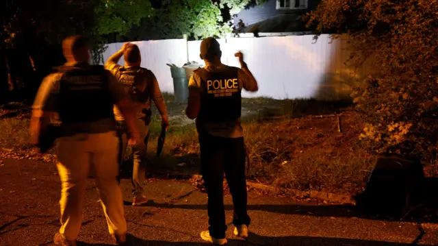 Three men, one wearing a police vest, shine torches on a fence near a suburban house