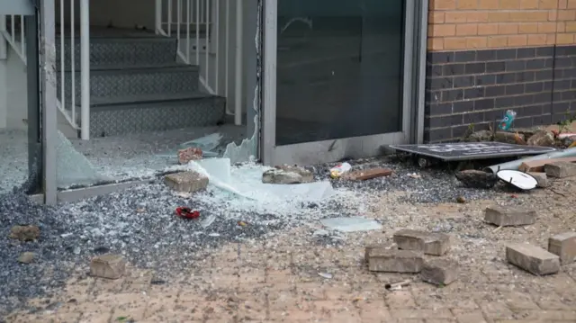 Shards of broken glass lie next to bricks and other objects outside the damaged window and door frame of a Holiday Inn Express hotel in Tamworth