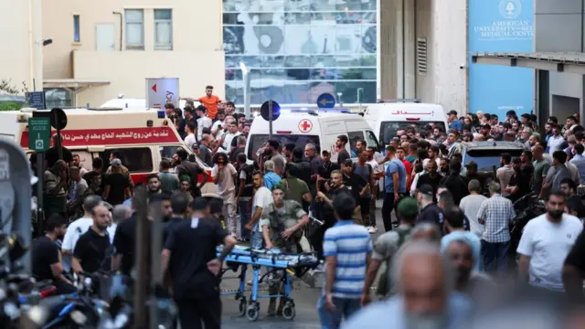 Ambulances line up outside a hospital, surrounded by a large crowd with men in uniform visible in the scene