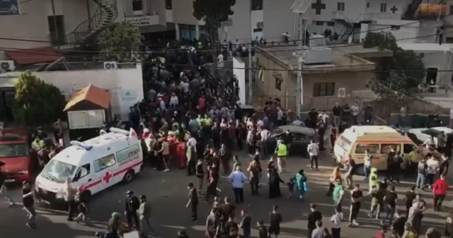 A crowd surrounds the open rear of a red cross van outside a building