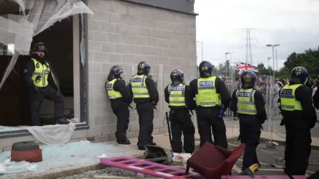 Line of police stand behind riot shields as one officer, wearing a helmet and body protection, emerges through a smashed window