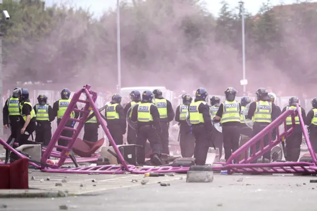 Line of police in riot gear with helmets and shields, with smoke drifting across and a series of upturned plastic barriers