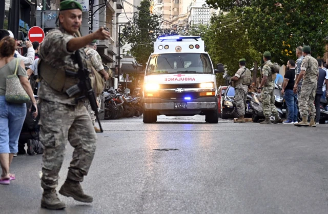 A soldier points at the forefront of a picture, waving a crowd away from the centre of the road as an ambulance makes its way towards his direction