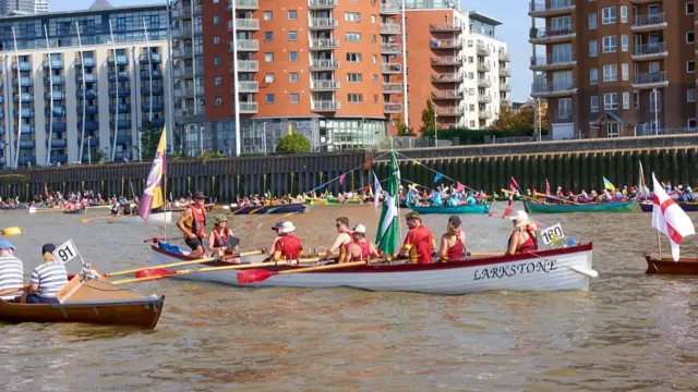 A boat adorned with bunting, with rowers wearing red and surrounded by other boats. Lettering on the side of the boat reads: Larkstone