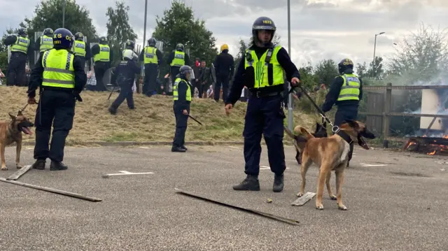 Police officers in riot gear with dogs on leashes surrounded by broken fence panels