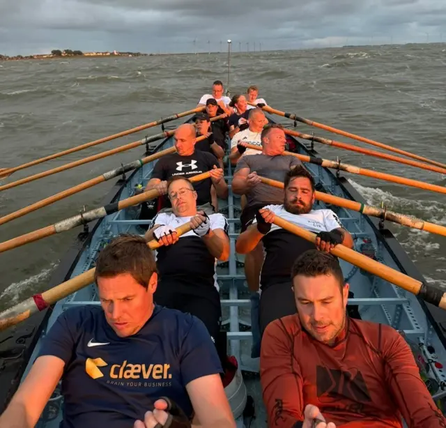 A picture of 12 people rowing, facing a camera and their faces looking as if it's a strenuous effort